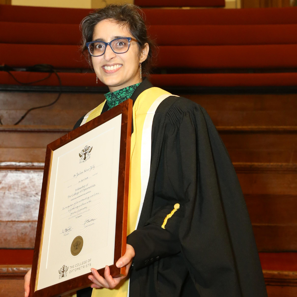 Dr Jolly is standing in front of wooden steps. She is dressed in a black academic gown with yellow and white trim, hair tied back, dark blue glasses, green turtle neck shirt and green earrings. Dr Jolly is holding a framed award certificate with a big smile.
